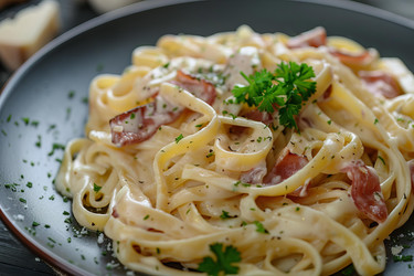 Fettuccine alfredo pasta dish is garnished with ham and parsley on black plate - yliv