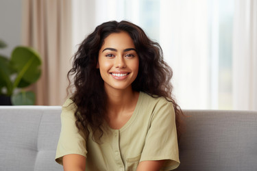 Portrait of a happy hispanic woman with curly hair sitting on a sofa in the living room and smiling - yliv
