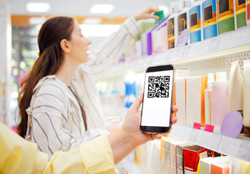 Покупательница держит в руках смартфон с QR-кодом, просматривая красочные косметические товары на полках в интерьере розничного магазина - Darya Petrenko