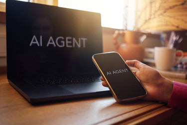 Смартфон с приложением AI Agent, который держит человек рядом с ноутбуком с таким же приложением на экране, деревянный стол с кофейной чашкой и растением на заднем плане - Darya Petrenko