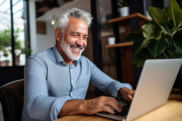Grey haired businessman smiling and typing on laptop, enjoying his work in a modern office - yliv