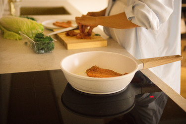 женщина-шеф-повар готовит филе лосося на деревянной разделочной доске на современной кухне со свежими овощами и белой сковородой на индукционной варочной панели - Darya Petrenko