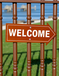 приветственный знак, размещенный на коричневом заборе, указывает прямо на зеленую лужайку. забор находится рядом с водоемом со спокойными волнами на заднем плане генеративный ии - Karjalainen