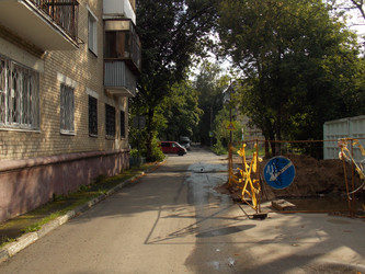 Городской двор с кирпичным жилым зданием слева, деревьями и участком дорожных или коммуникационных работ с оградительной лентой и дорожным знаком справа - Alenazam272