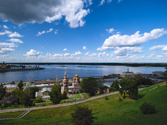 Вид на набережную Волги в Нижнем Новгороде. На переднем плане — зеленая холмистая местность с дорожками у Рождественской церкви. На заднем плане — река Волга, с островками и зелеными берегами. Вдали — городские здания и промышленные объекты. Небо ясное с  - Dimserg