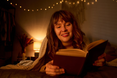 Улыбающаяся женщина читает книгу в уютной постели при теплом свете - Pressmaster