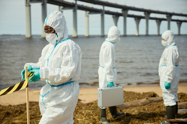 Чернокожая женщина в защитном костюме устанавливает предупреждающую ленту на загрязненном пляже - DragonImagesAsia