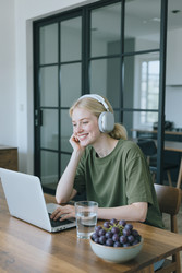 Улыбающаяся женщина в наушниках работает за ноутбуком в домашнем офисе - SeventyFour