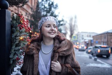 молодая женщина с рыжими волосами, одетая в меховую шубу и жемчужное ожерелье, стоит возле украшенного уличного столба, украшенного зимней зеленью и красными ягодами, в городской обстановке - Darya Petrenko
