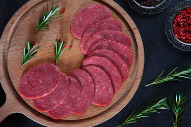нарезаем салями на деревянной доске, посыпаем розмарином и красным перцем. нарезанную колбасу салями разложите на деревянной разделочной доске. украсьте свежими веточками розмарина и горошинами красного перца. темный текстурированный фон. фотография мясно - RusAKphoto