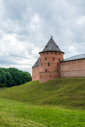 великий новгород, россия. башня метрополитен, массивная цилиндрическая крепостная башня из красного кирпича с крутой конической деревянной крышей, возвышающаяся над поросшим травой земляным валом под впечатляющим пасмурным небом - NikitaMaykov