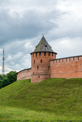 великий новгород, россия. федоровская башня новгородского кремля с круглой кирпичной крепостной стеной, конической крышей, средневековой оборонительной архитектурой, зеленой набережной и пасмурным летним небом - NikitaMaykov