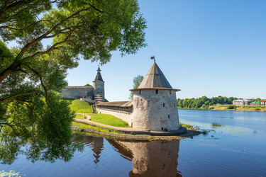 псков, россия. каменная плоская башня псковского крома, ее коническая крыша и грубые известняковые стены возвышаются над спокойными голубыми водами в месте слияния рек пскова и великая. - NikitaMaykov
