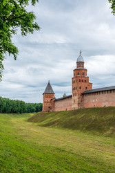 великий новгород, россия. стены и башни новгородского кремля из красного кирпича с высокой башней кокуй и ее характерной крышей, возвышающейся над заросшим травой рвом под пасмурным летним небом - NikitaMaykov