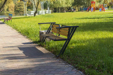 • Empty wooden bench swing on a paved park walkway next to a green lawn. Outdoors - Pazemin