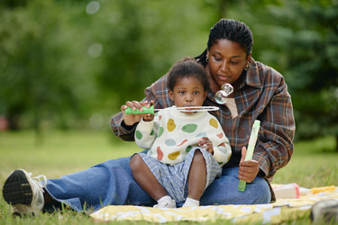 Чернокожая мать пускает мыльные пузыри с дочерью в парке - SeventyFour