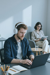Сосредоточенный мужчина в наушниках работает за ноутбуком в современном офисе - SeventyFour
