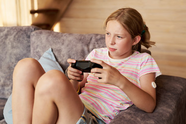Девушка с непрерывным монитором уровня глюкозы играет в видеоигру - Pressmaster